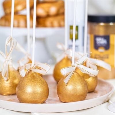 Bolinhos dourados com laço branco em palitos numa base branca.