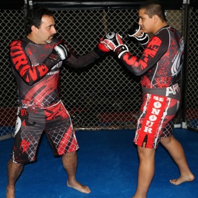 Dois homens a treinar MMA em ringue com chão azul e rede metálica ao fundo, usando roupa desportiva preta e vermelha com texto.