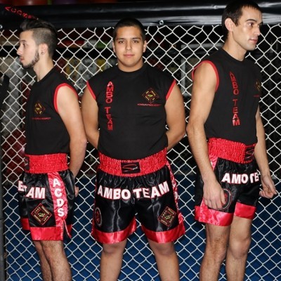 Três homens com roupas de treino pretas e vermelhas para artes marciais em fundo de rede metálica