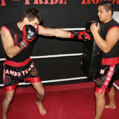 Dois homens a treinar boxe num ringue com calções pretos e vermelhos e equipamentos de treino