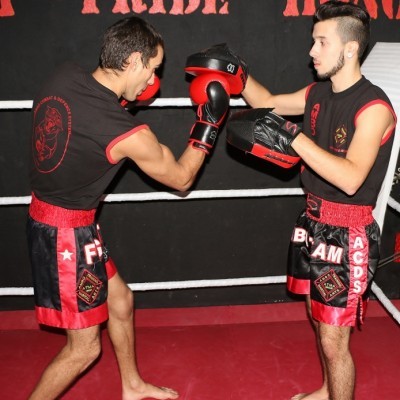 Dois homens em ringue de boxe com roupa de treino preta e vermelha, um com luvas de boxe vermelhas e outro com manoplas pretas.