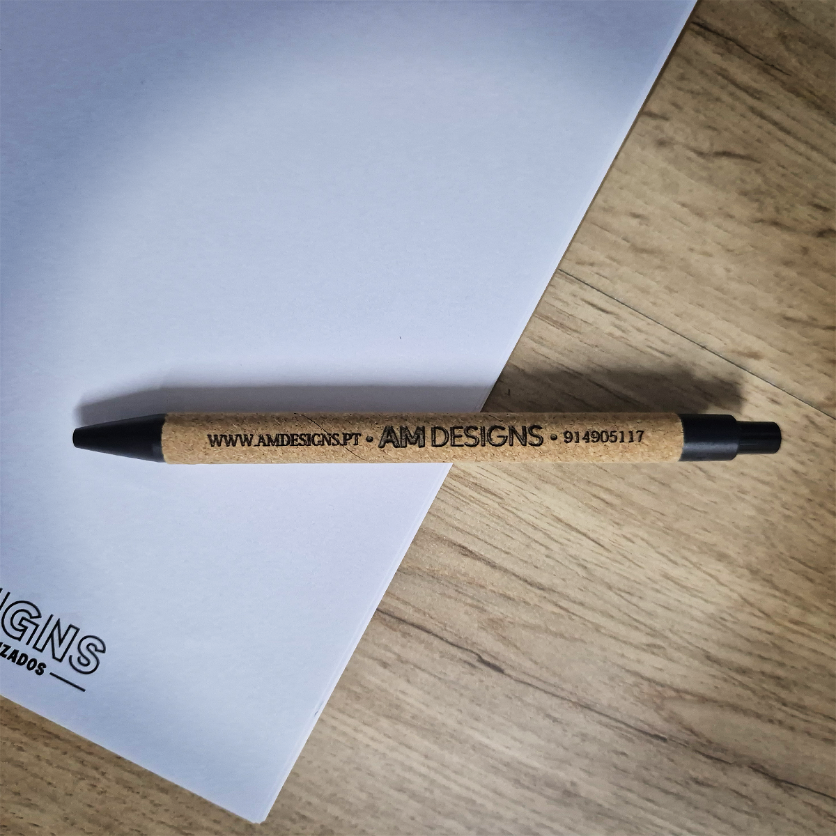 Brinde Empresarial – Caneta Eco Cork Personalizada Caneta de cortiça com texto preto sobre papel branco e superfície de madeira clara