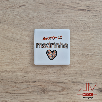 Quadrado branco decorativo com texto 'adoro-te madrinha' e coração floral sobre mesa de madeira