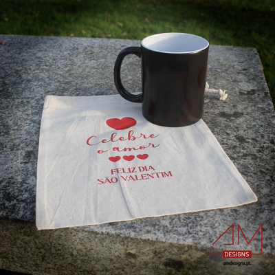 Caneca preta sobre saco de pano com texto em vermelho, sobre pedra, fundo de relva