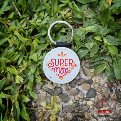 Chaveiro redondo branco com texto 'SUPER MÃE' em fundo natural