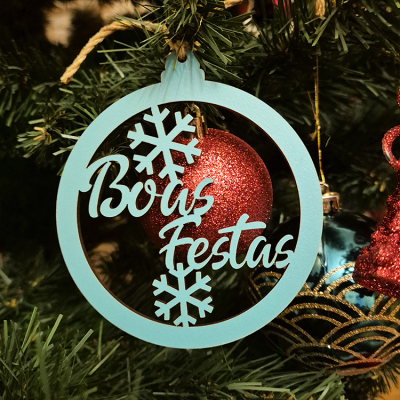 Adorno de Natal azul claro com texto 'Boas Festas' e flocos de neve pendurado em árvore com bola vermelha.