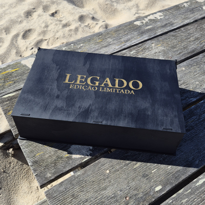 Caixa preta de madeira com texto dourado LEGADO EDIÇÃO LIMITADA numa mesa de madeira junto à areia