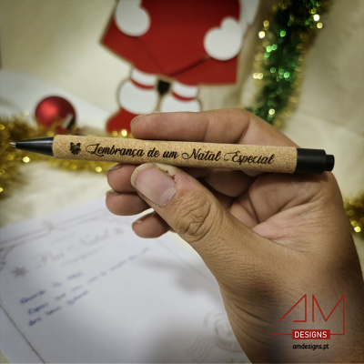 Caneta com texto 'Lembrança de um Natal Especial' segurada numa mão com decoração de Natal ao fundo