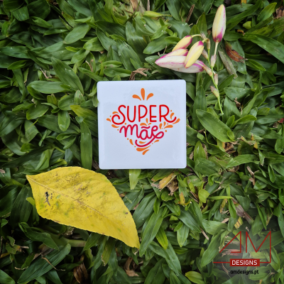 Caixinha branca com texto 'SUPER mãe' rodeada de folhas verdes e amarelas, botões de flor rosa, logo AM DESIGNS
