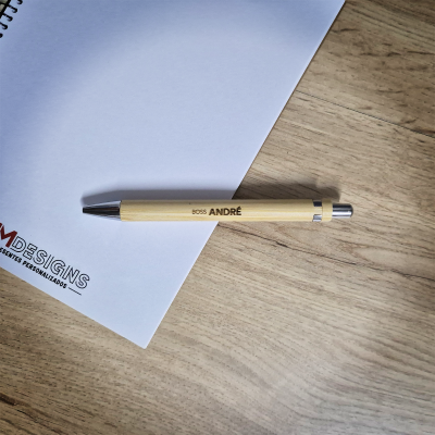 Caneta de madeira clara com texto 'BOSS ANDRÉ' ao lado de bloco de notas branco em espiral numa mesa de madeira clara.