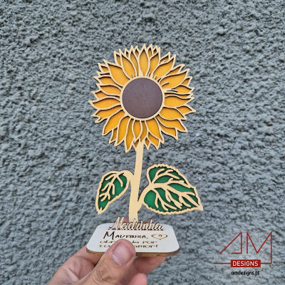 Escultura decorativa de girassol em madeira com base escrita, contra parede cinzenta.