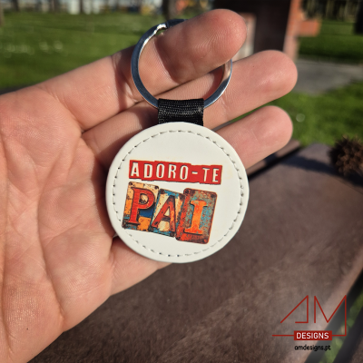 Chaveiro branco redondo com texto 'ADORO-TE PAI' segurado numa mão ao ar livre