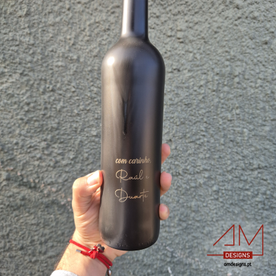 Garrafa de vinho preta fosca com texto dourado manuscrito segurada por uma mão contra uma parede cinzenta