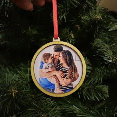 Ornamento de Natal redondo com foto de família e fita vermelha pendurado numa árvore.