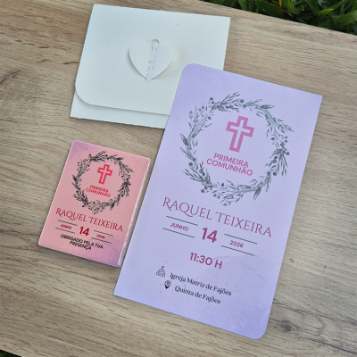 Convite e lembrança da Primeira Comunhão com envelope branco, convite roxo e lembrança rosa sobre mesa de madeira.