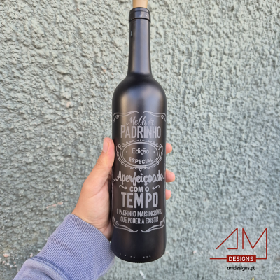 Garrafa preta com texto branco segurada por mão, fundo parede cinza