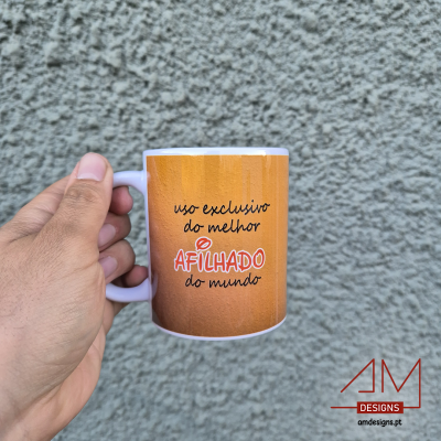 Caneca branca com mensagem para afilhado, fundo castanho, texto preto e vermelho, mão a segurá-la, parede cinza rugosa ao fundo.