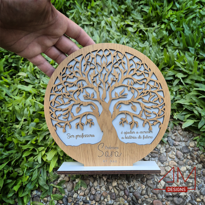 Suporte decorativo de madeira com árvore recortada e texto comemorativo para professora, mãos segurando, fundo de relva