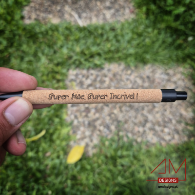 Caneta em cortiça com texto 'Super Mãe, Super Incrível!' segurada à frente de relva e caminho de pedras