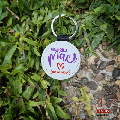 Chaveiro branco com texto MELHOR mãe DO MUNDO sobre relva e pedras