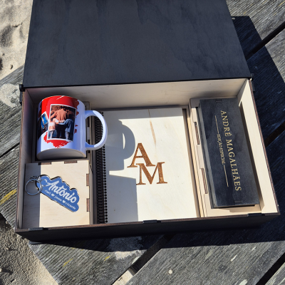 Caixa de presente com caneca personalizada, chaveiro, caderno de capa em madeira e caixa preta etiquetada, sobre madeira exterior.