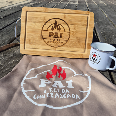 Conjunto para churrasco com tábua, caneca e avental com a inscrição PAI O REI DA CHURRASCADA e desenhos de fogo.