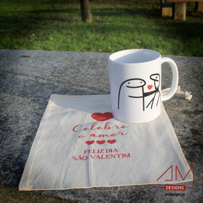 Caneca branca com figuras estilizadas e coração, em saco de pano com texto de Dia de São Valentim sobre pedra ao ar livre
