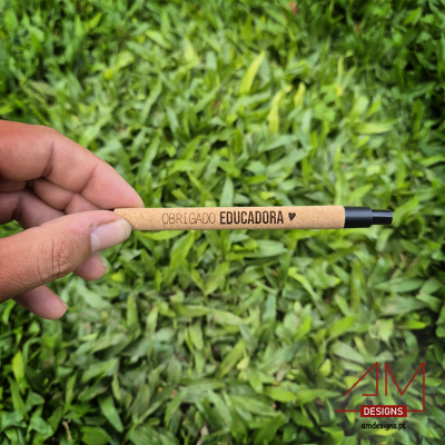 Caneta reciclada castanha clara com texto Obrigado Educadora, segurada sobre relva verde