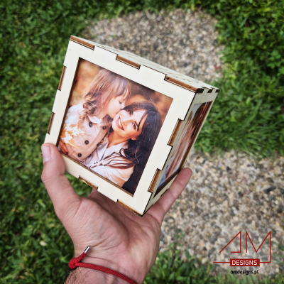 Caixa cúbica em madeira com fotos impressas em cada lado, segurada por uma mão, sobre calçada e relva