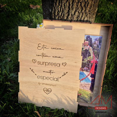 Caixa de madeira clara com texto gravado e puzzle colorido no interior junto a árvore.