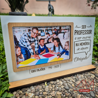 Moldura com foto de professor e crianças, texto de agradecimento em português, sobre base de madeira num espaço exterior.