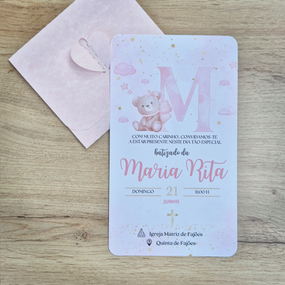 Convite de batizado com urso e estrelas, envelope cor-de-rosa, sobre mesa de madeira.