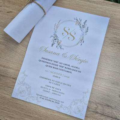 Convite de casamento branco texturizado com monograma dourado e detalhes florais ao lado de um rolo de papel amarrado com cordão