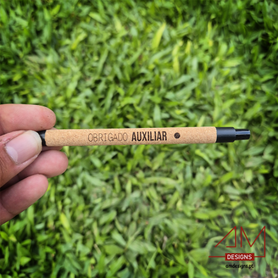Caneta castanha com texto 'OBRIGADO AUXILIAR' segurada em fundo de relva