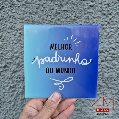 Azulejo azul com texto 'MELHOR padrinho DO MUNDO' segurado em frente a parede cinza.