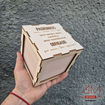Caixa de madeira clara com texto gravado para padrinhos, segurada por uma mão em fundo cinza.