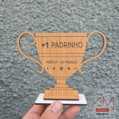 Troféu de madeira claro com base branca, inscrição #1 PADRINHO melhor do mundo, contra fundo cinzento.