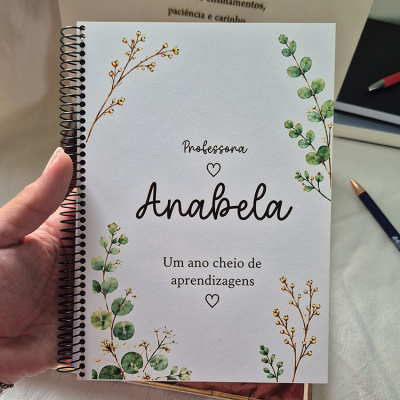 Caderno espiral branco decorado com ramos verdes e amarelos e texto dedicado a professora