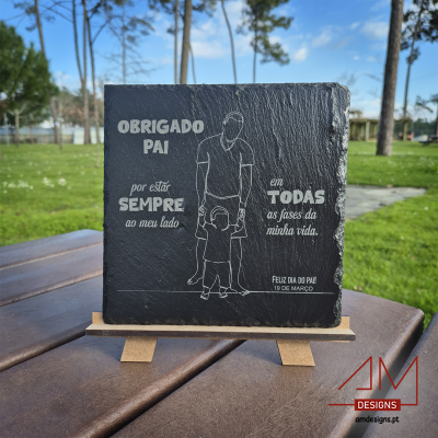 Placa decorativa em ardósia preta com texto e ilustração de pai e filho, em cavalete sobre mesa ao ar livre