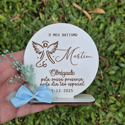 Placa redonda decorativa branca com texto e laço azul, sobre base castanha, segurada por mão em fundo verde