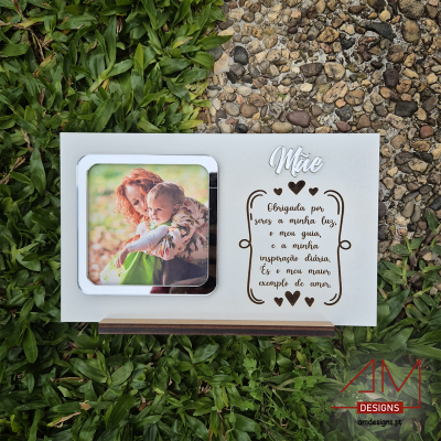 Painel branco com porta-fotos metálico e texto gravado para mãe, exposto sobre relva e pedra