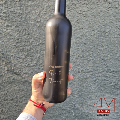 Garrafa preta fosca com texto dourado segura por mão em frente a parede cinza e logo AM DESIGNS