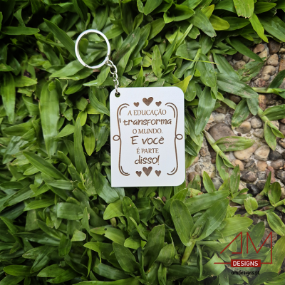 Porta-chaves de madeira branca com texto gravado sobre fundo de folhas verdes e pedrinhas