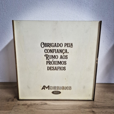 Caixa quadrada de madeira clara com texto gravado a preto de agradecimento e logotipo AM DESIGNS 2025.
