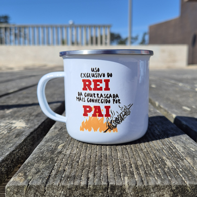 Caneca metálica branca com texto e imagem de fogo e espetada, em cima de mesa de madeira ao ar livre