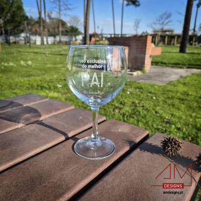 Copo de vidro para vinho com texto alusivo ao pai, em ambiente de parque com mesa de madeira
