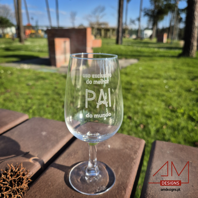 Copo de vidro com texto gravado 'uso exclusivo do melhor PAI do mundo' sobre mesa de madeira ao ar livre