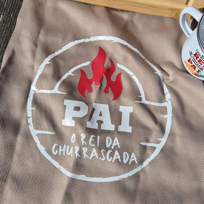 Avental bege com estampa PAI O REI DA CHURRASCADA e chama vermelha, sobre superfície de madeira com caneca branca ao lado