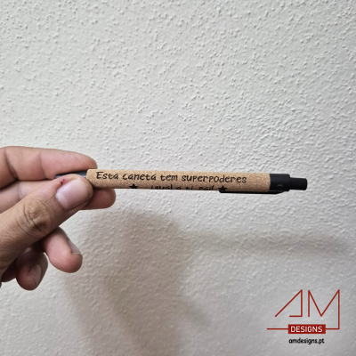 Caneta com corpo em cortiça clara e texto 'Esta caneta tem SUPERPODERES', ponta preta e botão de clique preto, segurada por mão contra parede branca