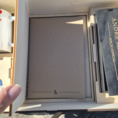 Caixa de madeira aberta com caderno castanho, livro preto e caneca branca com desenho vermelho